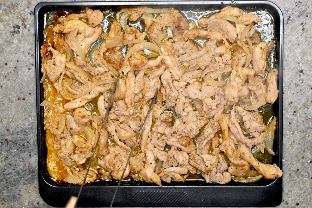Roasted chicken shawarma strips with onions on a sheet pan with tongs.