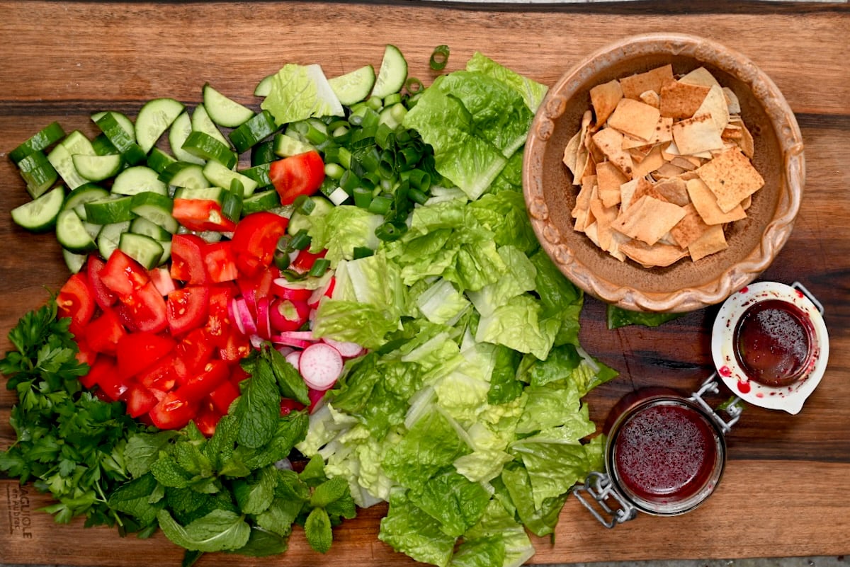 Fattoush salad ingredients on a cutting board with chopped romaine, cucumber, tomato, radish, herbs, pita chips, and sumac dressing.
