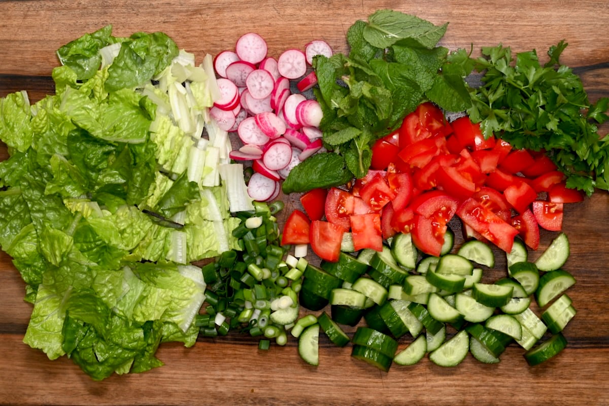 Prepped fattoush vegetables on a cutting board with chopped romaine, cucumber, tomato, radish, green onion, mint, and parsley.
