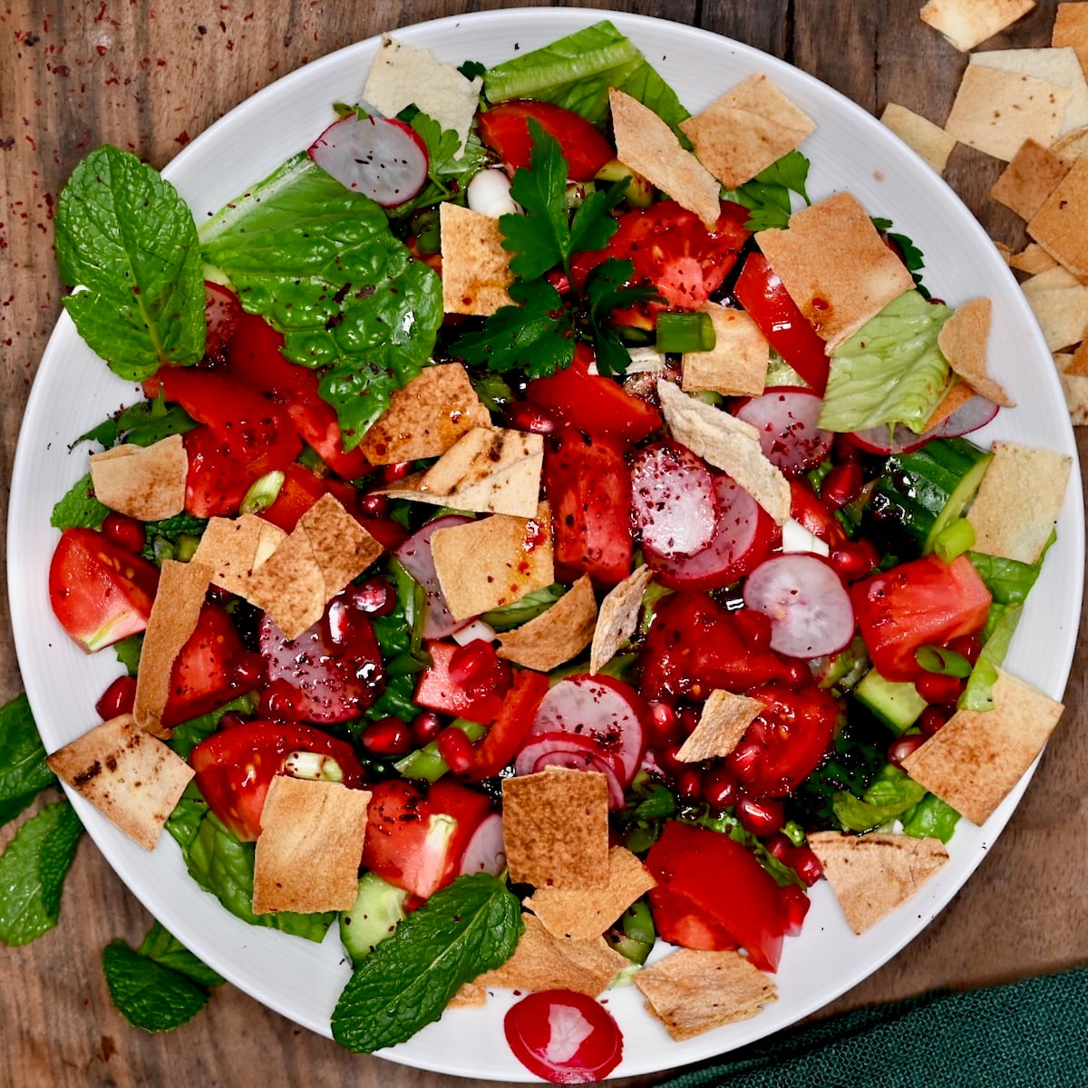 Fattoush salad on a plate with romaine, tomatoes, cucumber, radish slices, pomegranate seeds, pita chips, and sumac dressing drizzle.