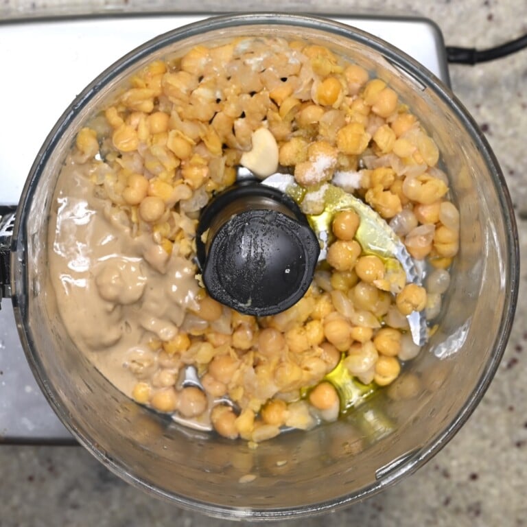 Food processor bowl with chickpeas, tahini, olive oil, garlic, and salt before blending.