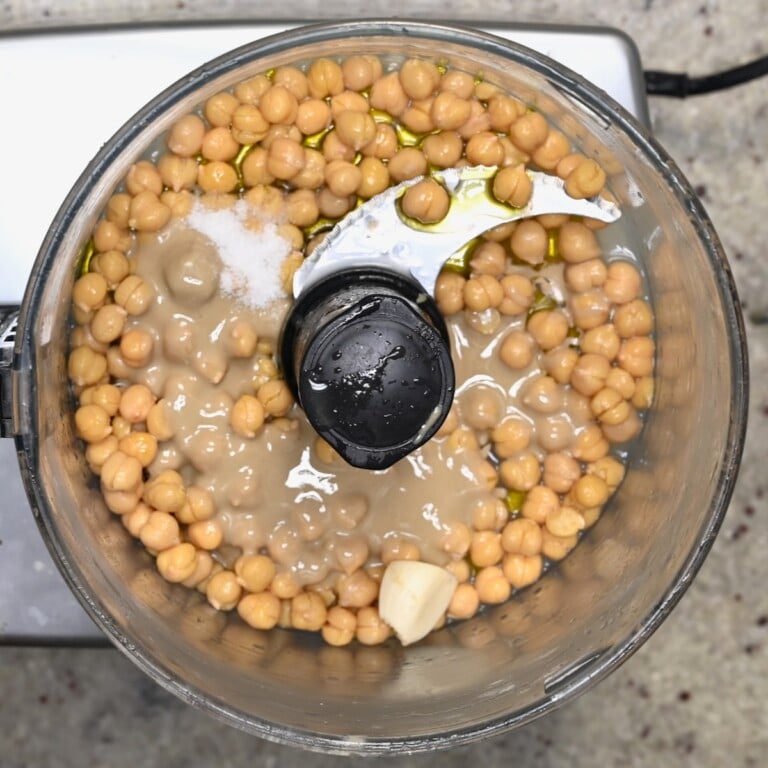 Food processor bowl with chickpeas, tahini, olive oil, garlic, and salt before blending.