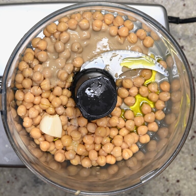 Food processor bowl with chickpeas, tahini, olive oil, and a garlic clove before blending.