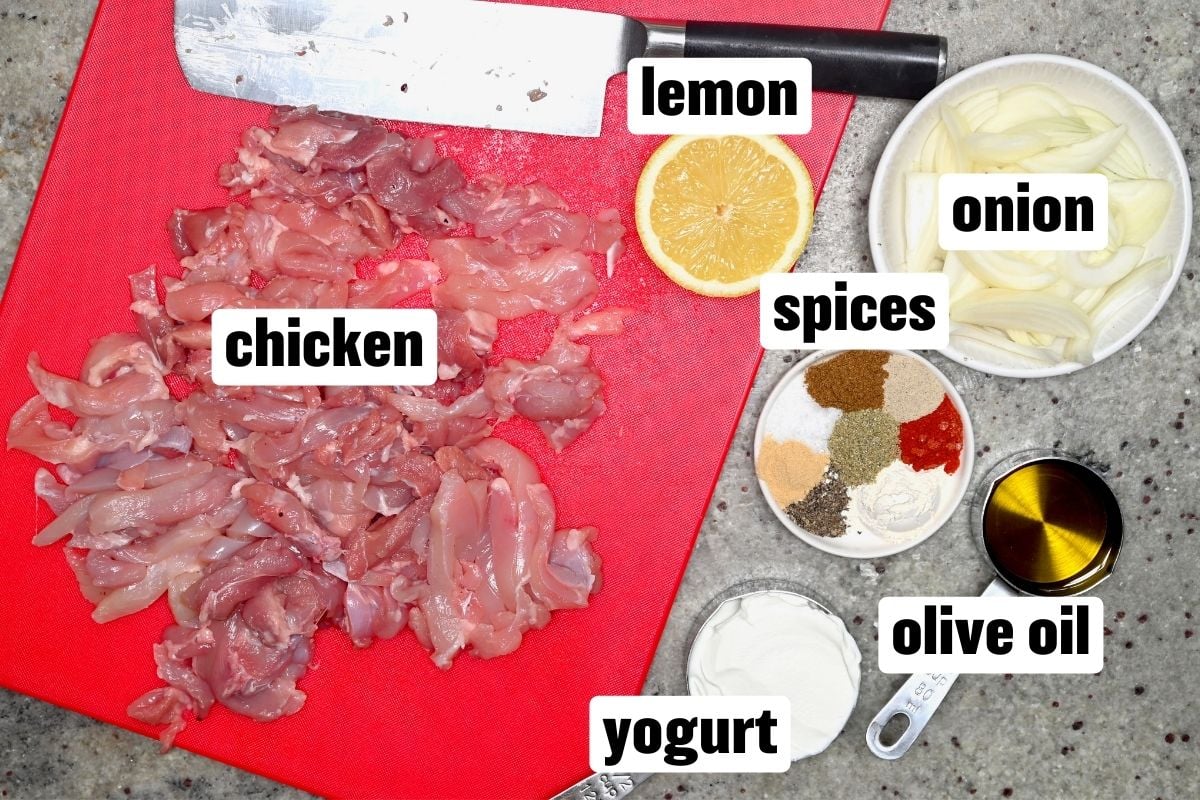 Ingredients for chicken shawarma on a cutting board with sliced chicken, lemon, onion, yogurt, olive oil, and mixed spices