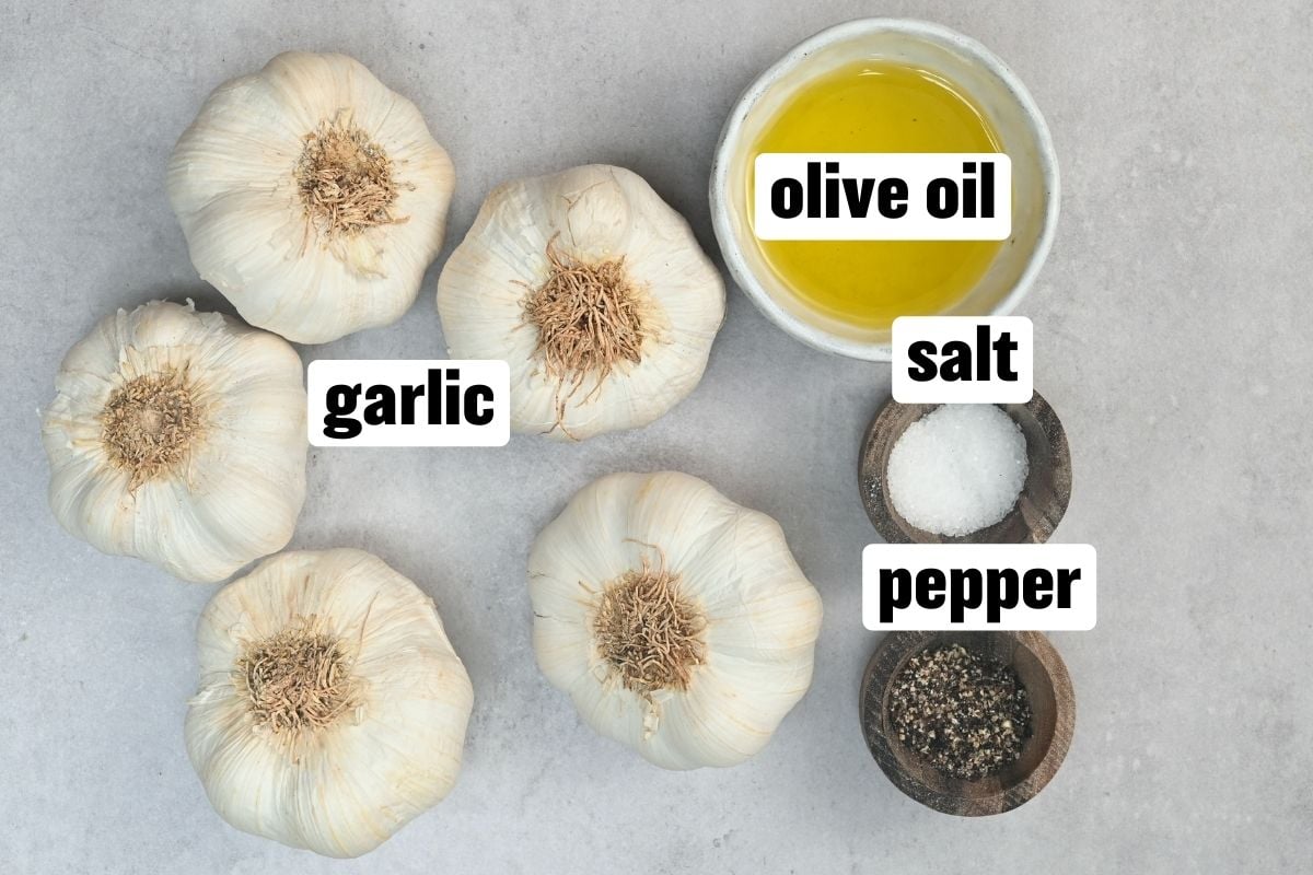Flat lay of whole garlic bulbs with small bowls of olive oil, salt, and black pepper on a light gray surface.