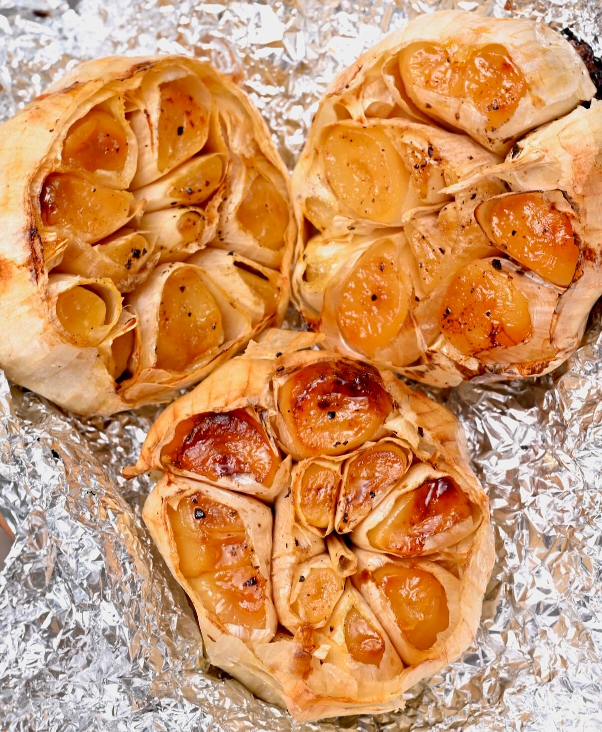 Three roasted garlic heads in foil with soft, golden caramelized cloves.