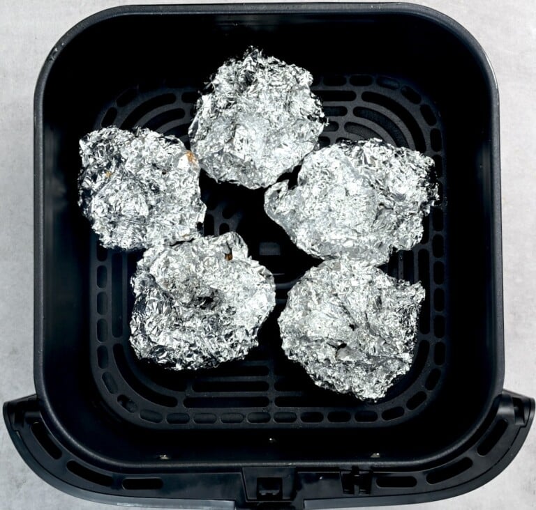 Air fryer basket filled with foil-wrapped garlic heads ready to roast.