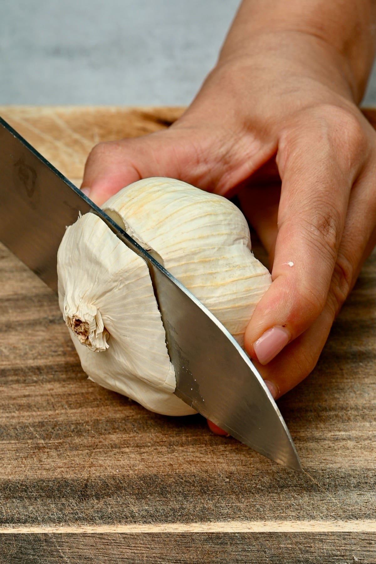 Knife slicing the top off a whole garlic bulb on a wooden cutting board.