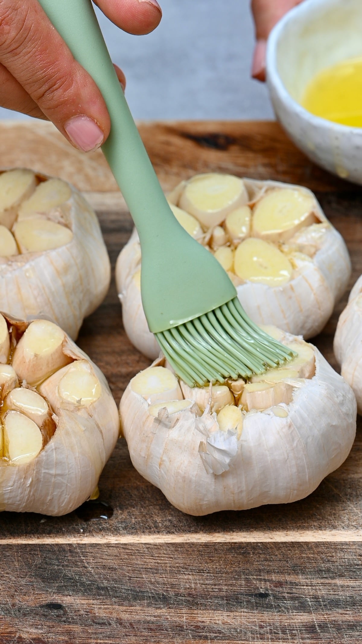 Silicone brush coating cut garlic heads with olive oil on a wooden board.