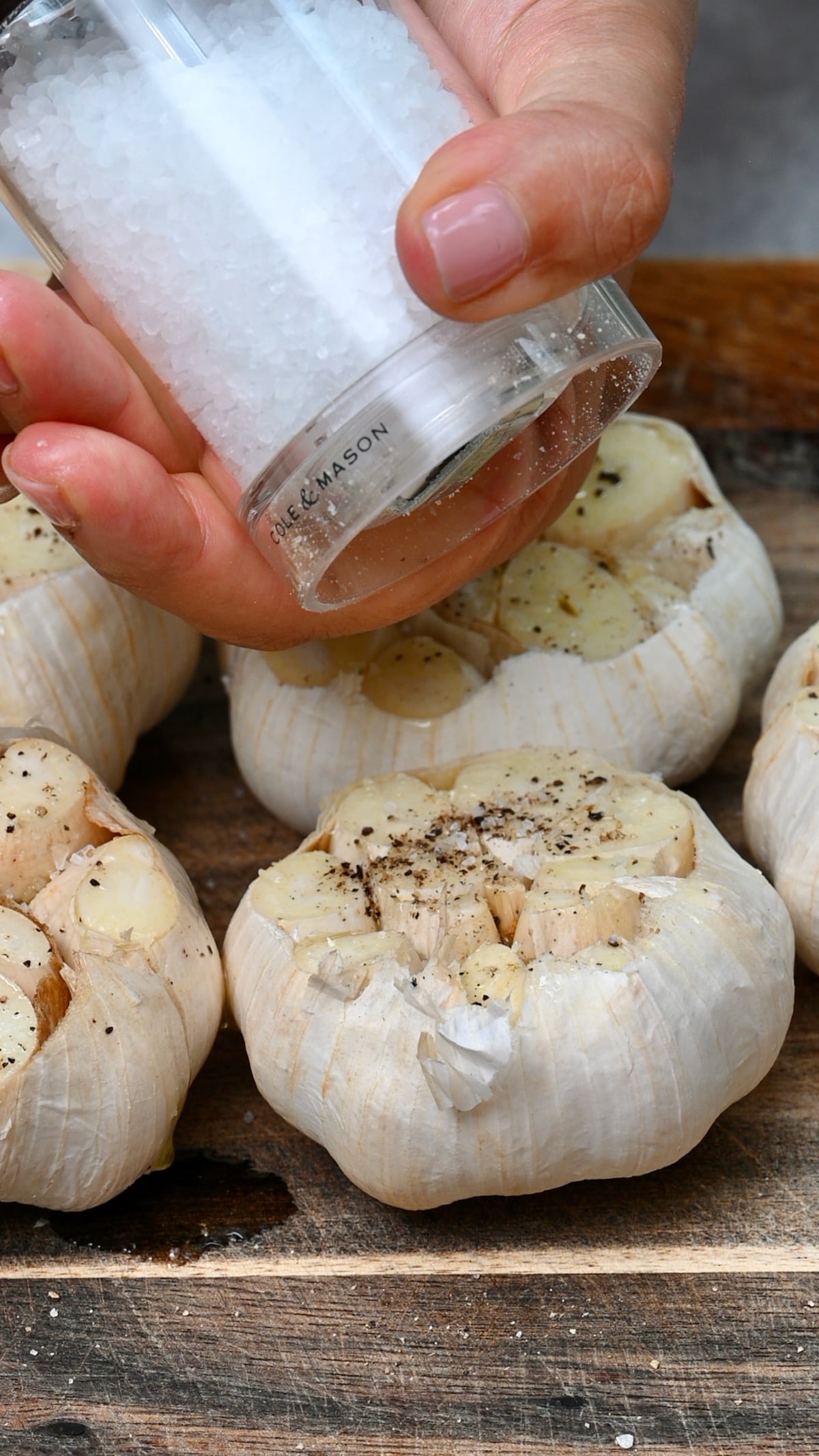 Hand sprinkling salt over cut garlic heads seasoned with black pepper on a wooden board.