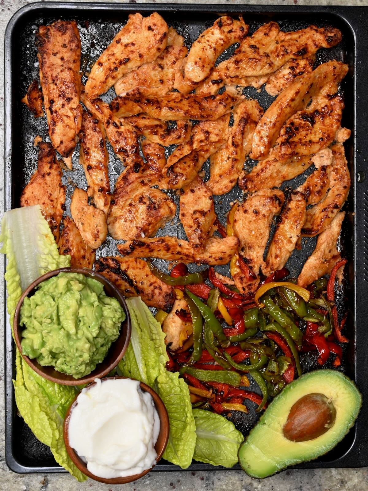 Baked chicken tenders on a tray with lettuce leaves, fajita peppers, guacamole, garlic sauce, and half an avocado.