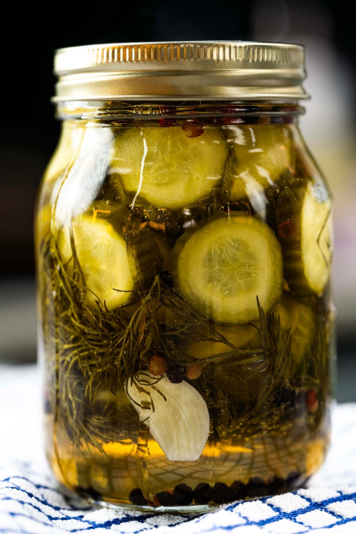 Jar of homemade dill pickle slices with fresh dill, garlic, and peppercorns in brine, shown close-up on a kitchen towel.