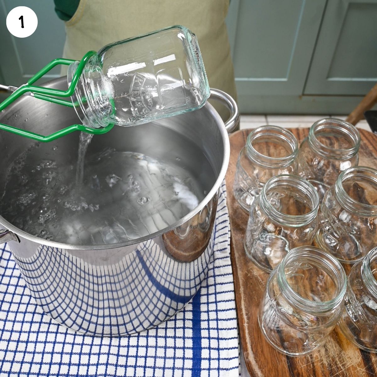 Glass jars being sterilized in boiling water, with one jar lifted from a large pot and more empty jars arranged on a wooden board.