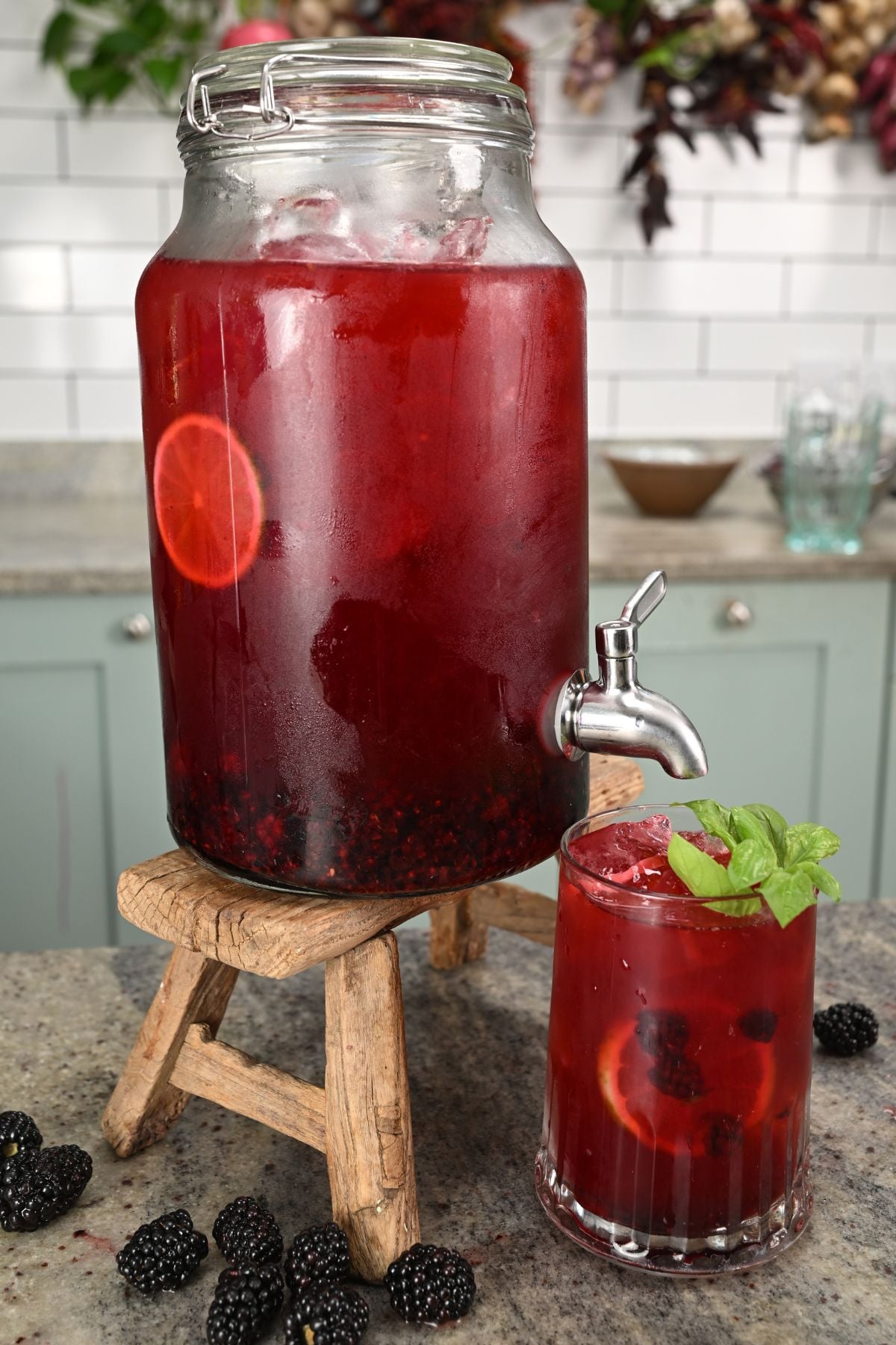 A glass drink dispenser with a spigot sitting on a small wooden stand, filled with iced red blackberry drink, next to a filled glass garnished with fresh basil leaves, lemon slices, and blackberries on a countertop.