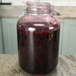 A large glass latch-top jar filled with dark purple macerated blackberry syrup, sitting on a kitchen countertop.