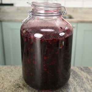 A large glass latch-top jar filled with dark purple macerated blackberry syrup, sitting on a kitchen countertop.