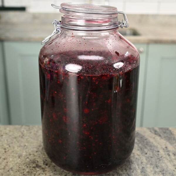 A large glass latch-top jar filled with dark purple macerated blackberry syrup, sitting on a kitchen countertop.