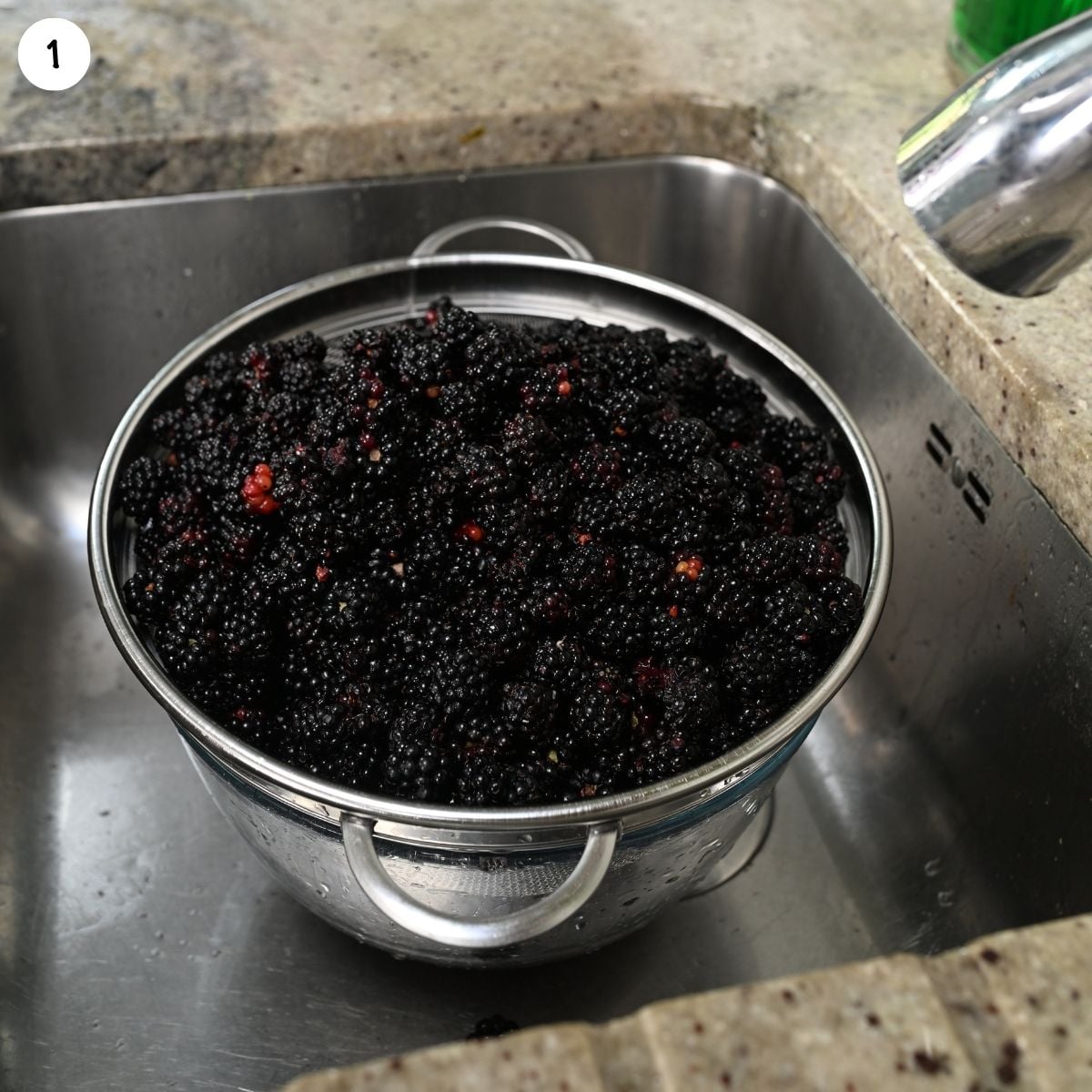 A stainless steel colander filled with fresh, dark blackberries sitting inside a kitchen sink after being washed.