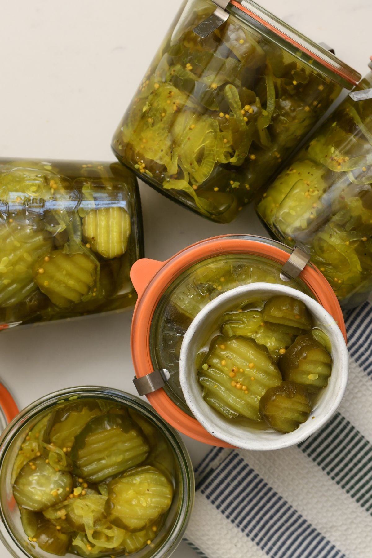 Overhead view of glass Weck jars filled with bread and butter pickles alongside a small ceramic bowl of pickle slices on a striped towel.