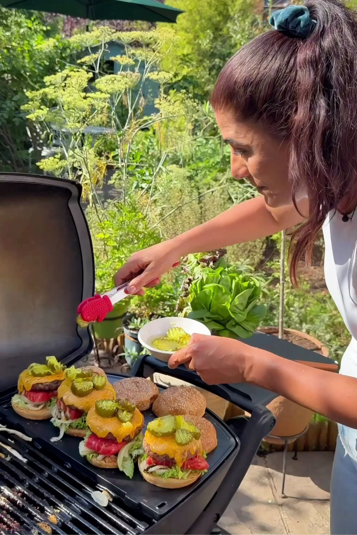 Samira standing outdoors at an open grill, using red tongs to add bread and butter pickles from a small white bowl onto grilled cheeseburgers lined up on the grate.