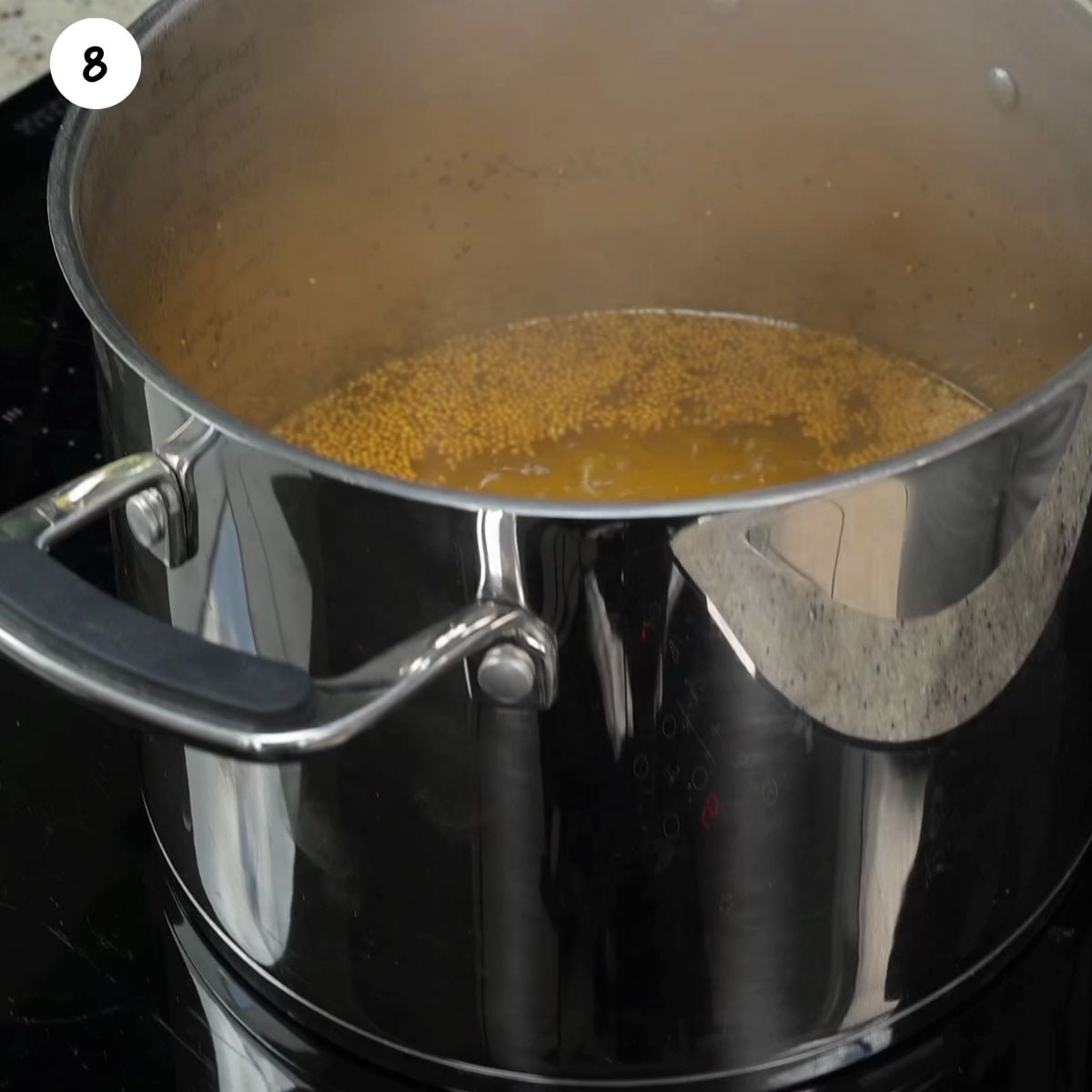 A large stainless steel pot on a black stovetop filled with a steaming, golden-yellow pickling brine containing visible mustard seeds.