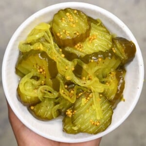 Top-down view of a hand holding a small white bowl filled with sweet and tangy crinkle-cut bread and butter pickles.