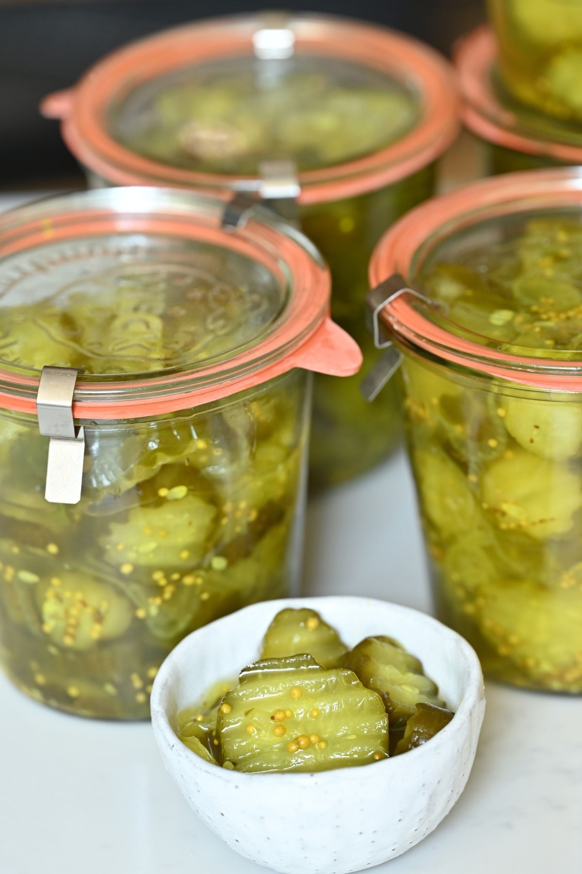 Close-up of a small white bowl with crinkle-cut bread and butter pickles, set in front of glass Weck jars filled with the pickles.