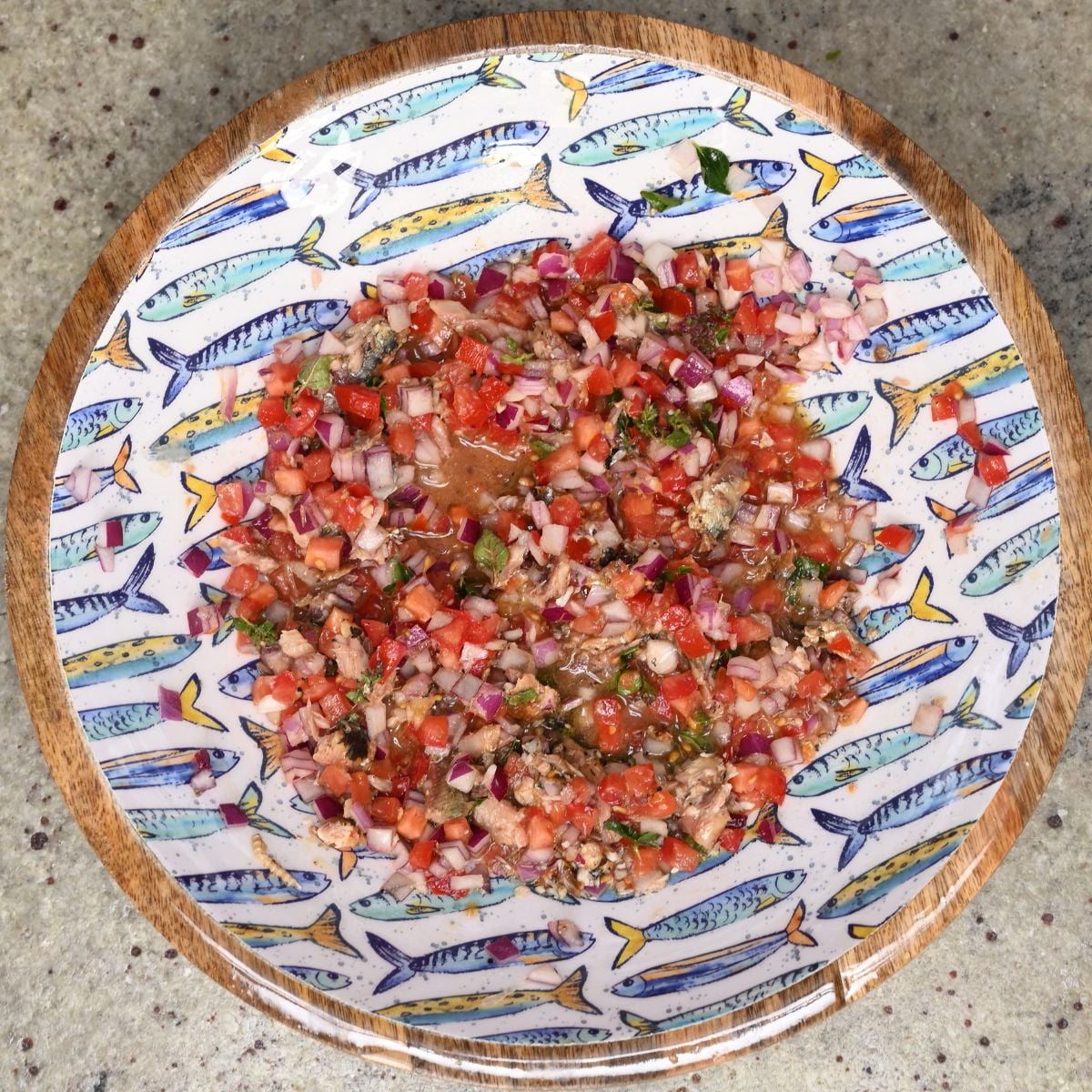 Top-down view of a fresh canned sardine salad mixed with diced tomatoes and red onions, served in a decorative round plate with a blue fish pattern.