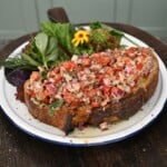A thick slice of toasted sourdough bread generously topped with a sardine, tomato, and red onion salad, served on a plate alongside fresh mixed greens.