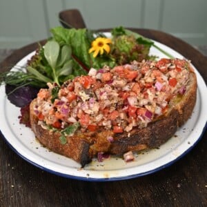 A thick slice of toasted sourdough bread generously topped with a sardine, tomato, and red onion salad, served on a plate alongside fresh mixed greens.
