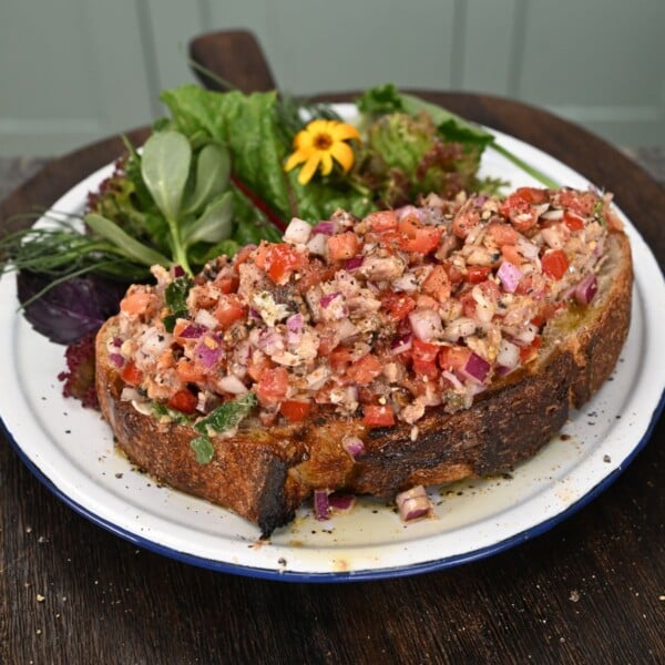 A thick slice of toasted sourdough bread generously topped with a sardine, tomato, and red onion salad, served on a plate alongside fresh mixed greens.