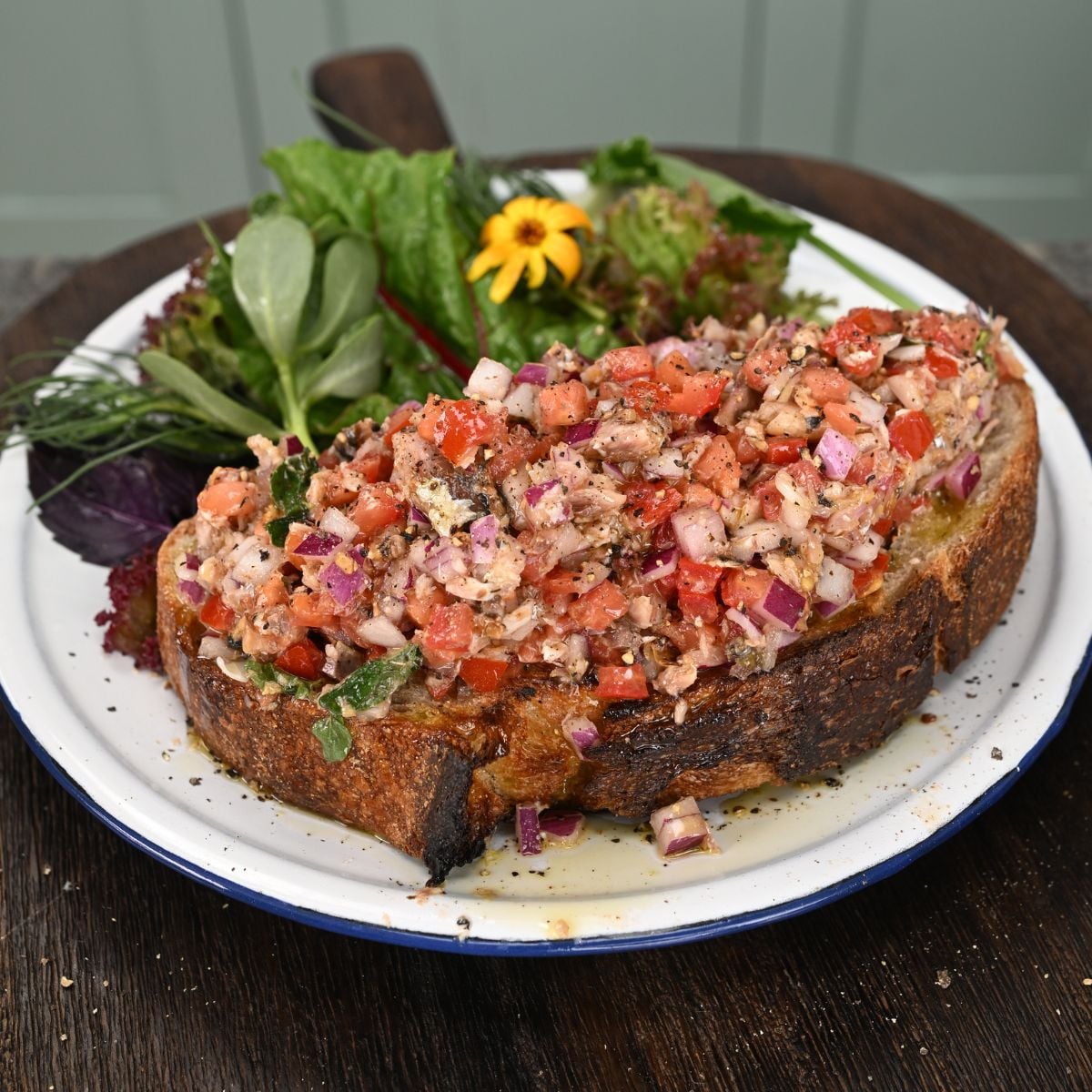 A thick slice of toasted sourdough bread generously topped with a sardine, tomato, and red onion salad, served on a plate alongside fresh mixed greens.