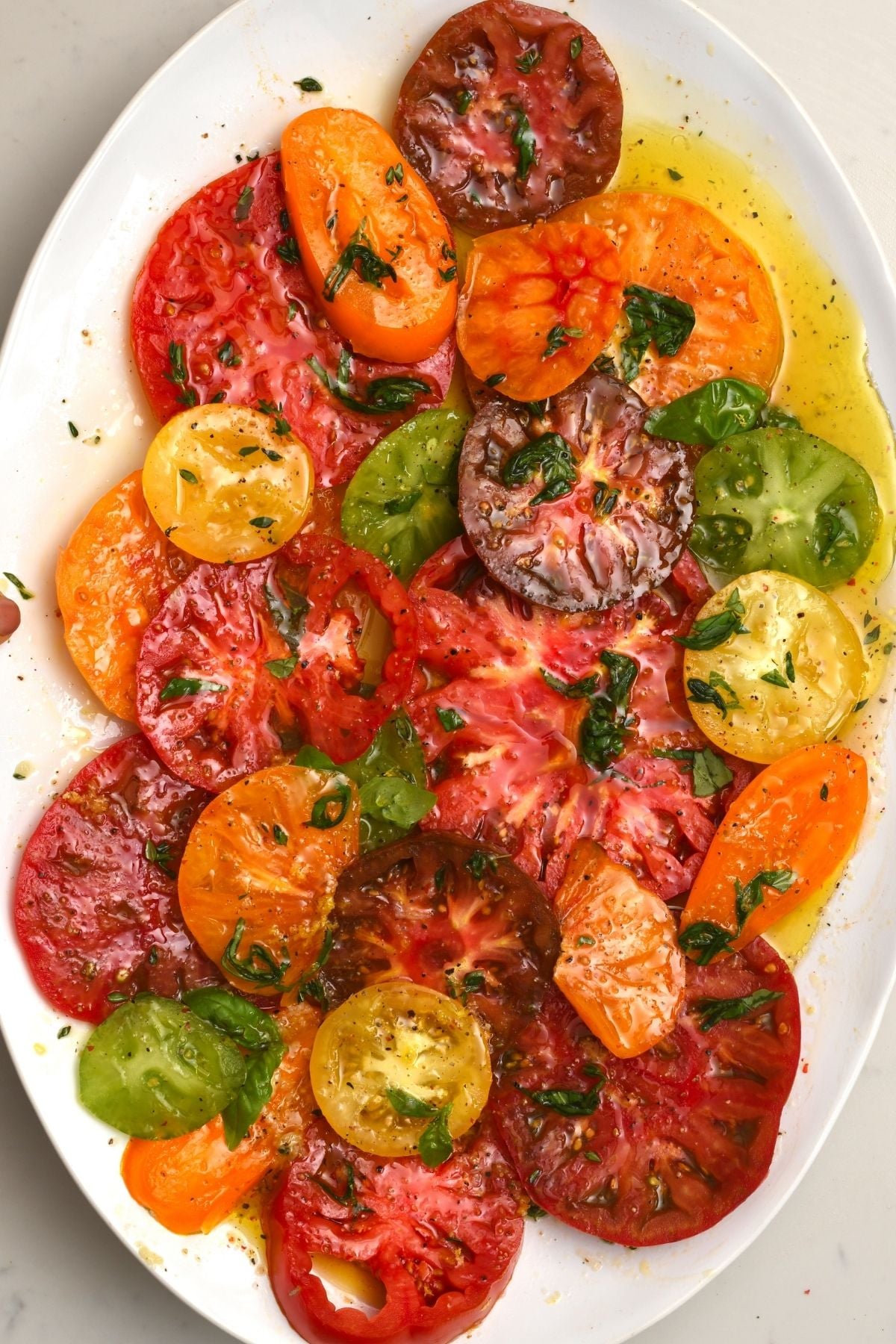 Slices of red, yellow, orange, green, and dark purple heirloom tomatoes arranged on a white oval platter, dressed with olive oil, black pepper, and fresh basil.