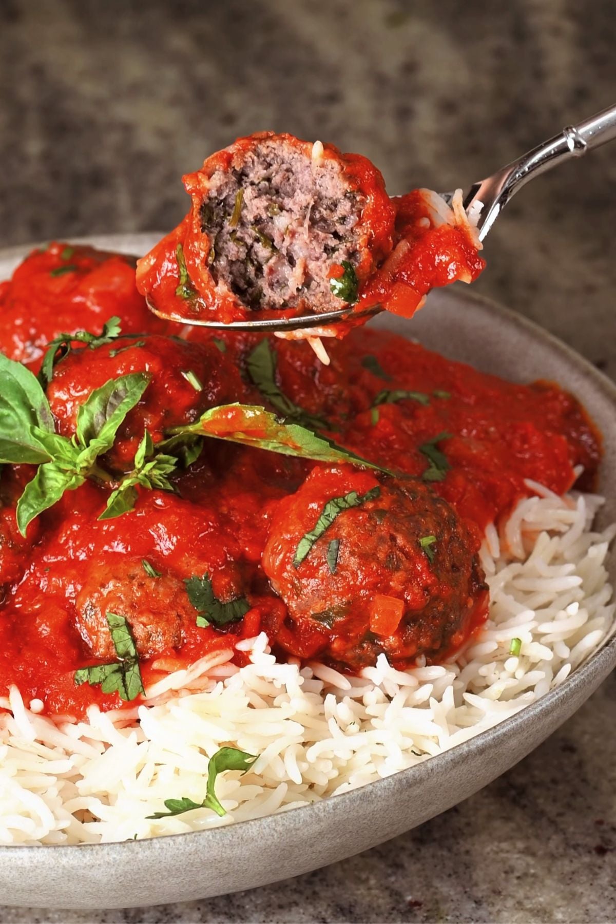 A fork holding a half-eaten kofta meatball coated in rich red tomato sauce over a bowl of long-grain white rice and kofta meatballs garnish with fresh herbs.