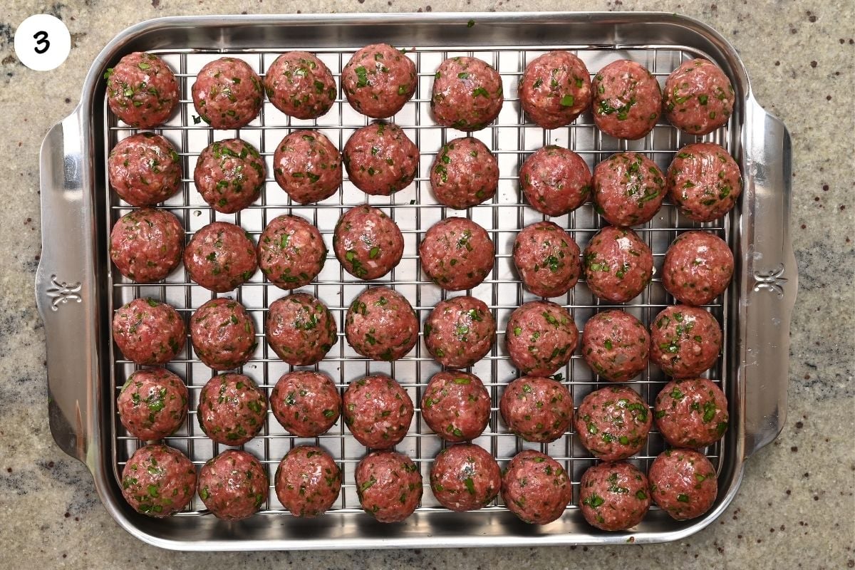 Top-down view of raw, hand-rolled kofta meatballs with visible chopped parsley, arranged neatly in rows on a wire rack inside a stainless steel baking tray.