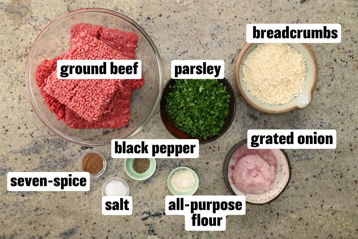 Top-down view of ingredients for kofta meatballs arranged on a countertop, including ground beef, breadcrumbs, parsley, grated onion, flour, seven-spice, black pepper, and salt.