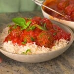A bowl of fluffy white basmati rice topped with savory kofta meatballs drenched in vibrant tomato sauce, garnished with fresh basil and parsley leaves, with a cooking pot of meatballs visible in the background.