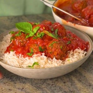 A bowl of fluffy white basmati rice topped with savory kofta meatballs drenched in vibrant tomato sauce, garnished with fresh basil and parsley leaves, with a cooking pot of meatballs visible in the background.