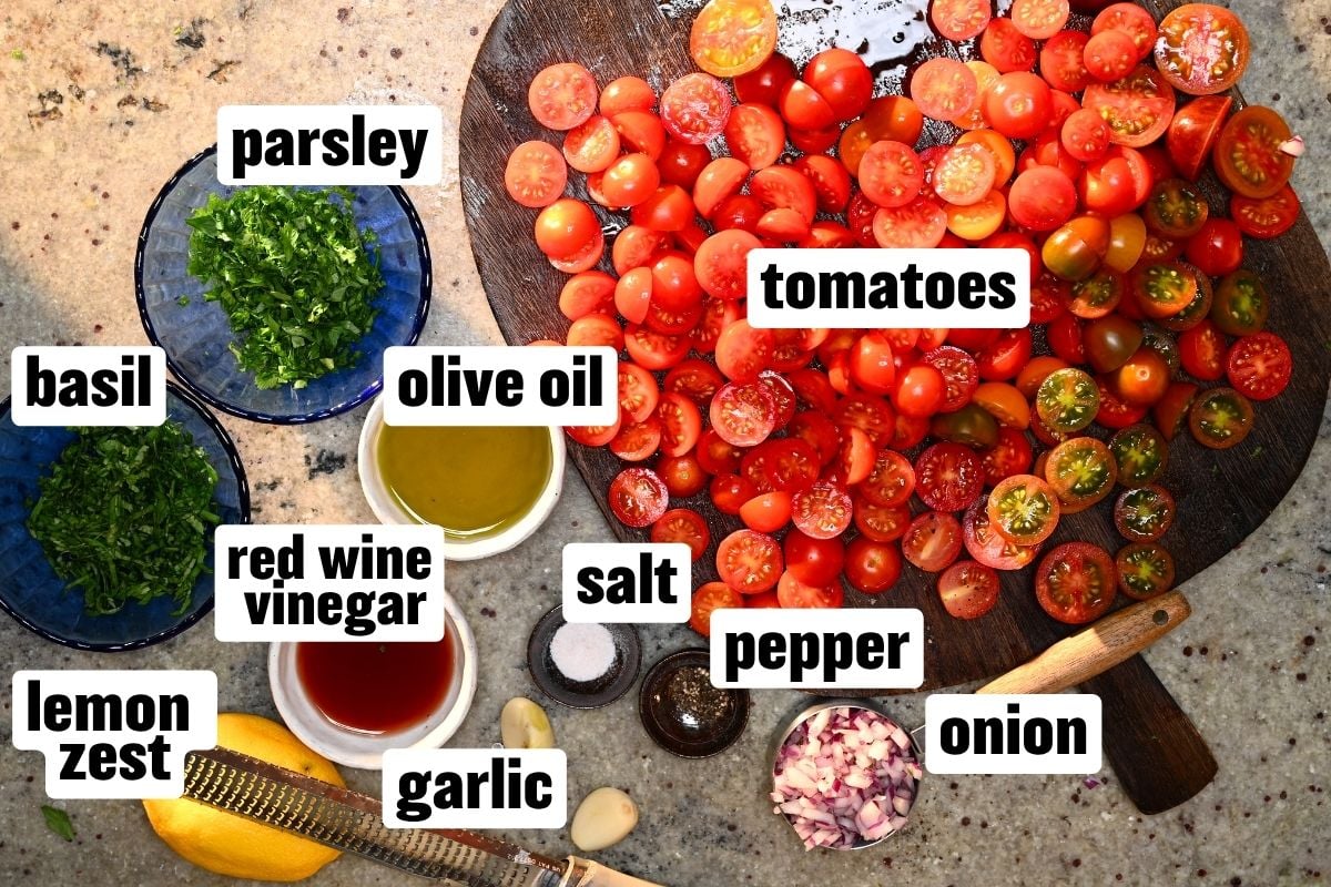 Sliced cherry tomatoes on a wooden cutting board with bowls of chopped parsley, basil, diced onion, olive oil, red wine vinegar, garlic, lemon zest, salt, and pepper.
