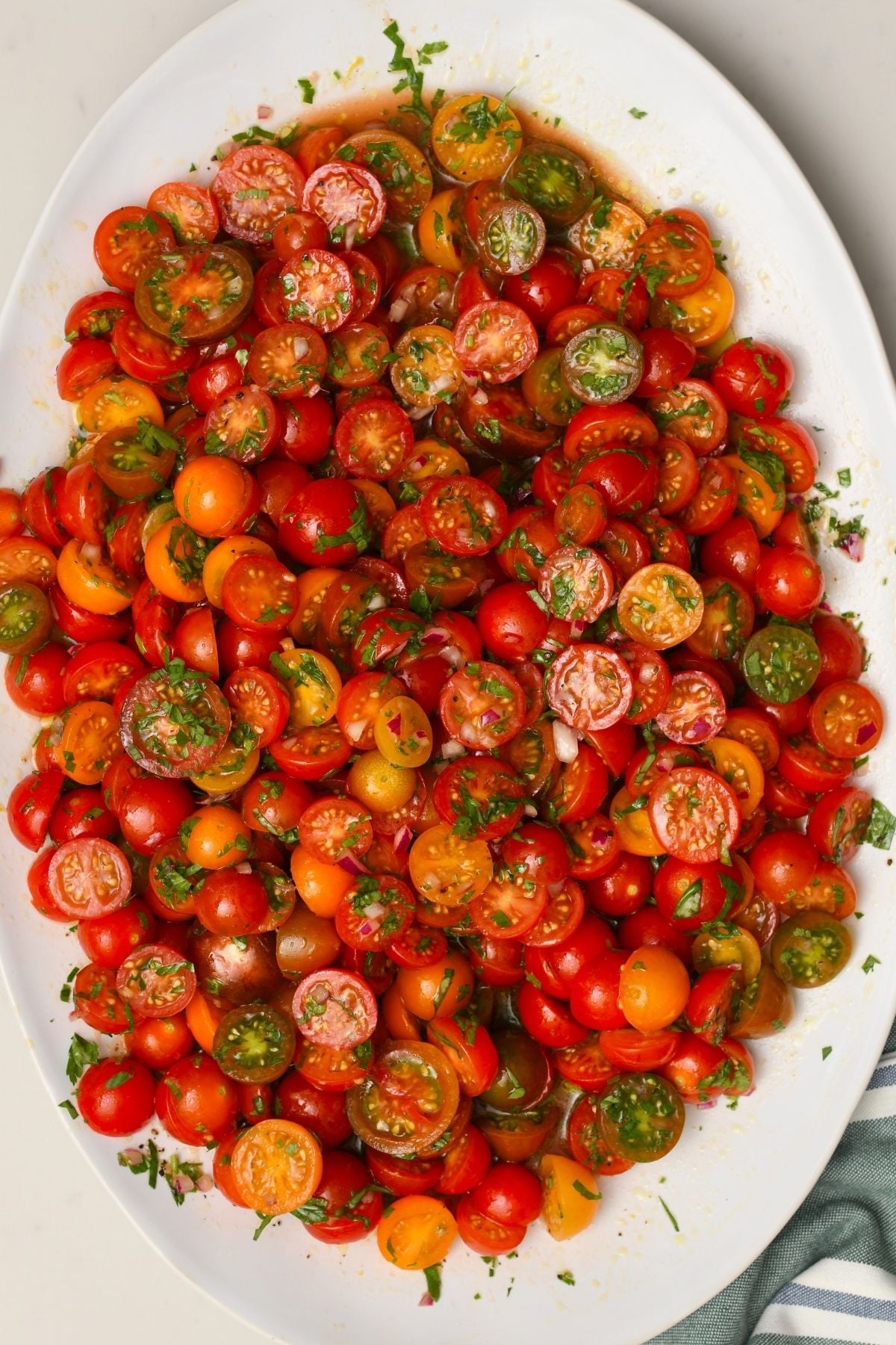 A large white oval platter filled with halved red, orange, and green cherry tomatoes tossed in fresh herbs and marinade.