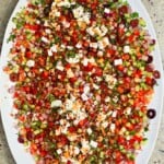 Close-up top-down view of a large platter filled with colorful Mediterranean olive oil dip, loaded with finely diced tomatoes, cucumbers, red onion, Kalamata olives, crumbled feta cheese, and fresh herbs.