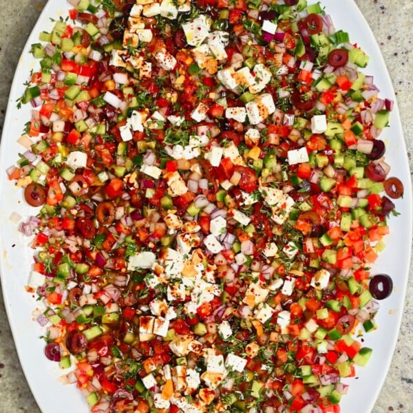 Close-up top-down view of a large platter filled with colorful Mediterranean olive oil dip, loaded with finely diced tomatoes, cucumbers, red onion, Kalamata olives, crumbled feta cheese, and fresh herbs.