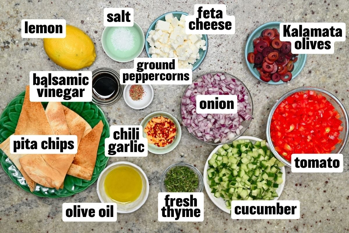 Flat lay view of ingredients including diced tomatoes, cucumbers, red onion, feta cheese, Kalamata olives, pita chips, olive oil, lemon, balsamic vinegar, fresh thyme, and spices arranged on a countertop.