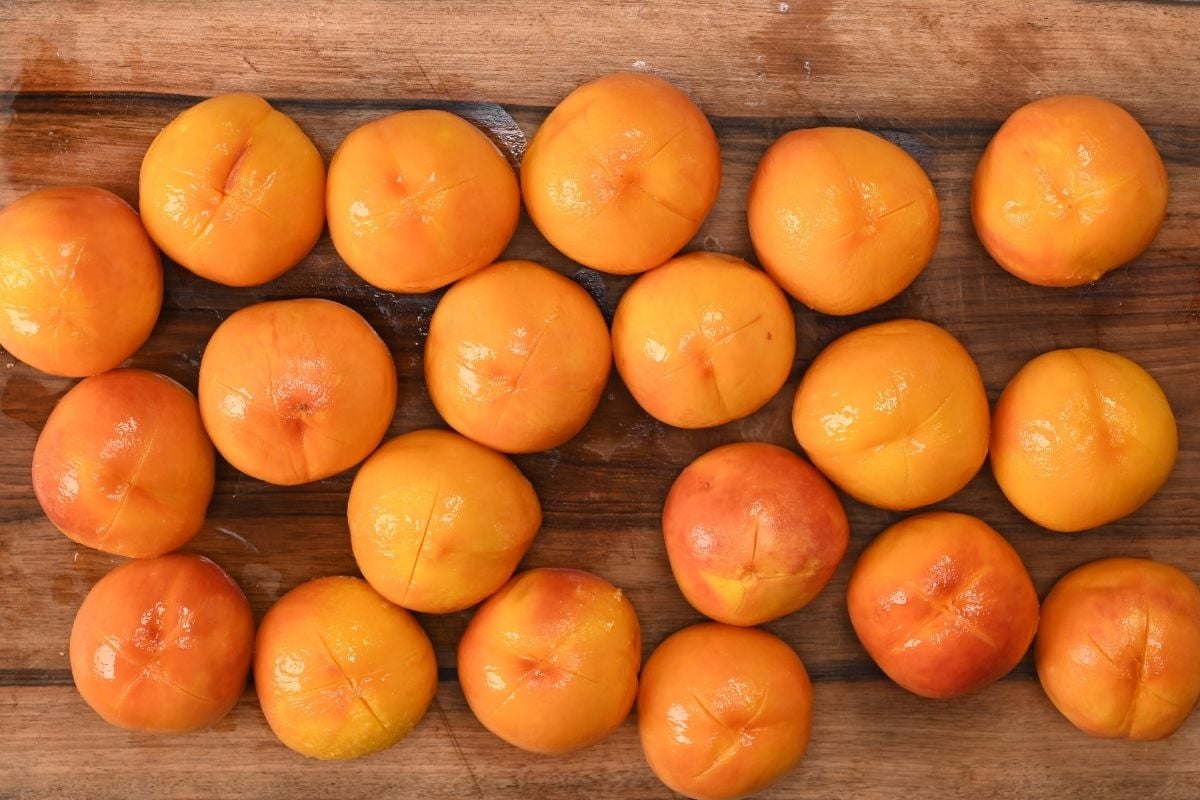 Top-down view of whole blanched and peeled fresh peaches marked with scored crosses on the bottom, arranged across a wooden cutting board.