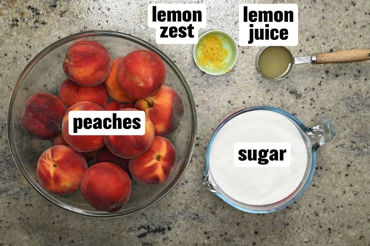 Top-down view of ingredients for peach butter arranged on a countertop, including a glass bowl of fresh whole peaches, a measuring cup of sugar, a small bowl of lemon zest, and lemon juice.