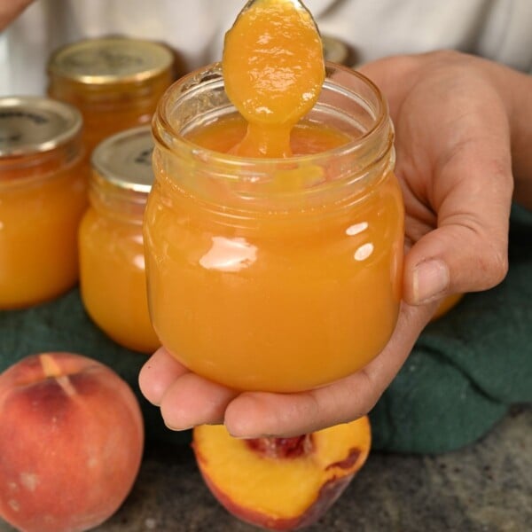 Close-up of a hand holding an open glass jar of smooth, golden peach butter with a spoon lifting out a spoonful, set against additional sealed jars and fresh peaches.