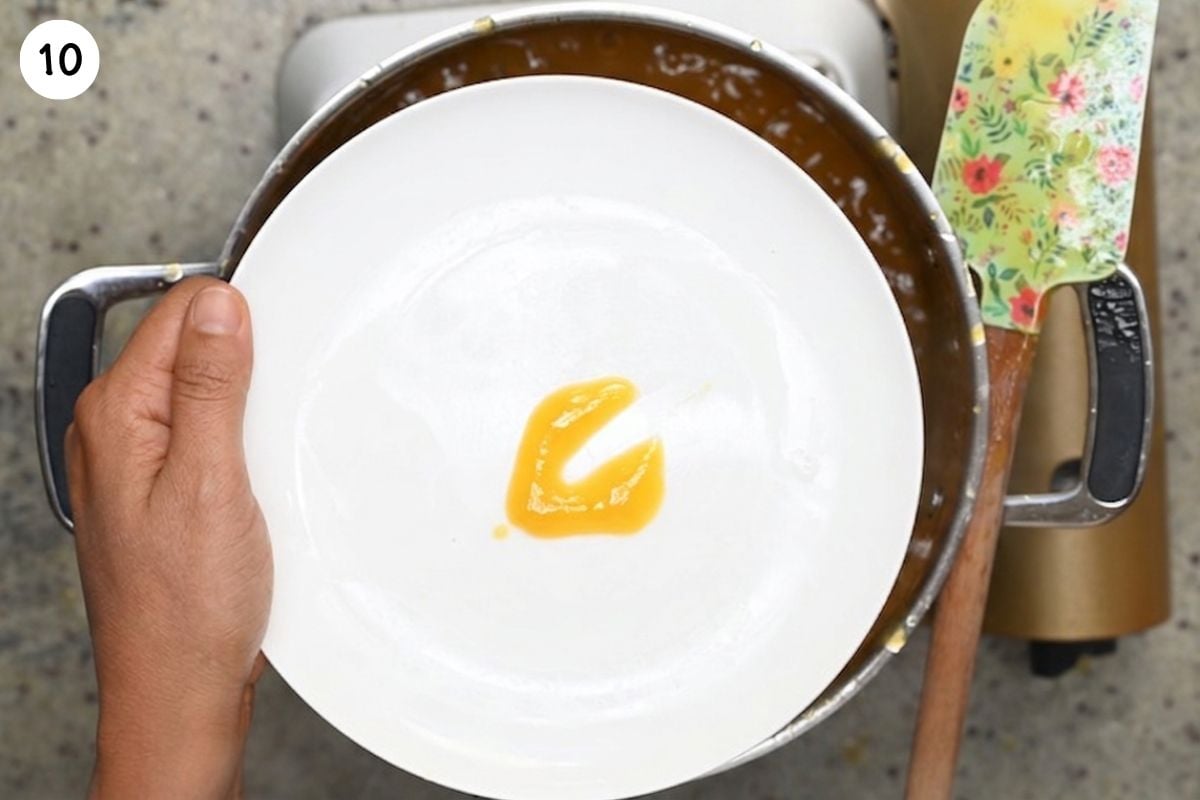 Top-down view of a hand holding a chilled white plate with a spoonful of cooked peach butter, featuring a clear line dragged through it to test for proper set and gel consistency.
