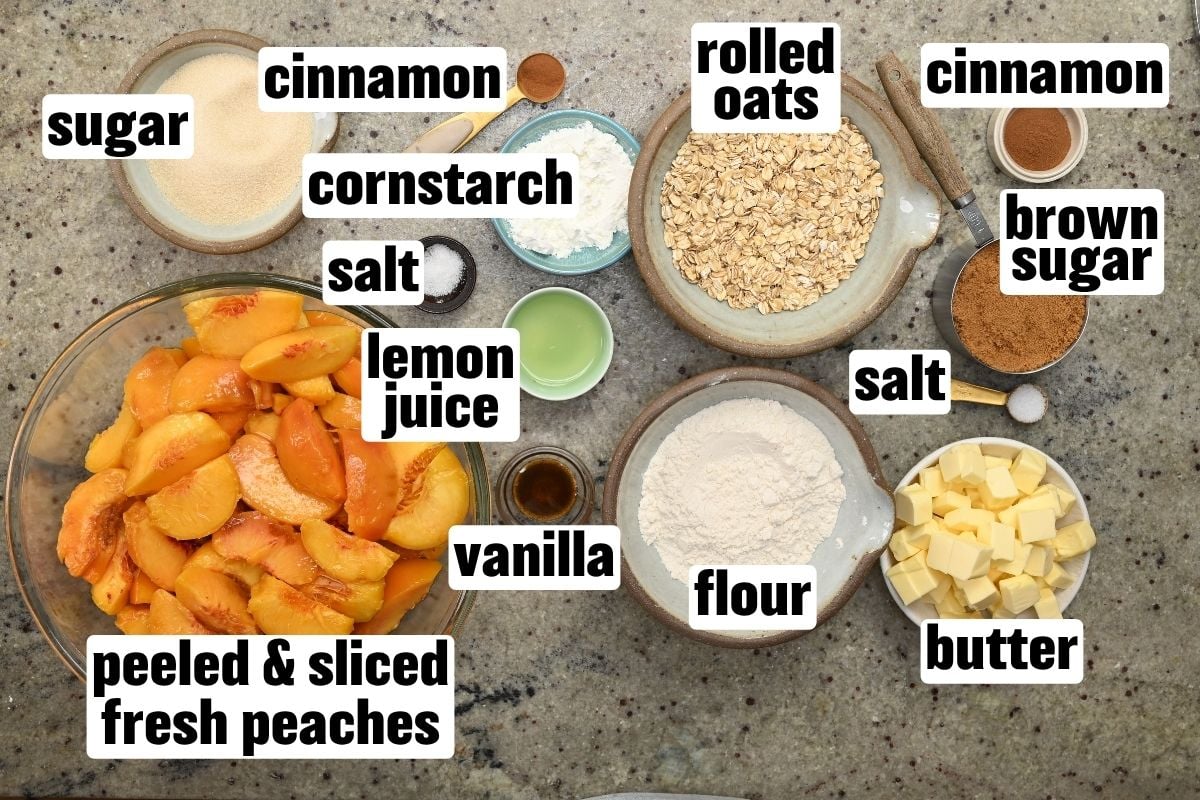 Top-down view of peach crisp ingredients, including sliced fresh peaches, sugar, brown sugar, rolled oats, flour, butter, cornstarch, lemon juice, vanilla, salt, and cinnamon arranged in bowls on a countertop.