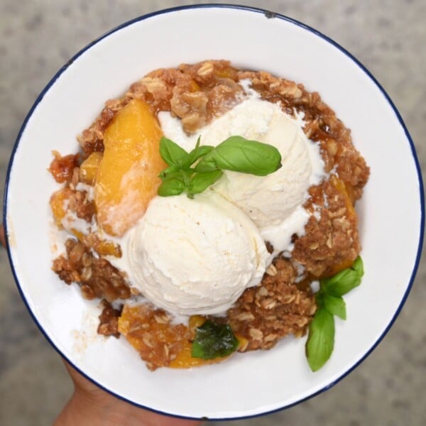 Top-down view of a hand holding a white enamel plate filled with warm peach crisp topped with two scoops of vanilla ice cream and fresh basil sprigs.