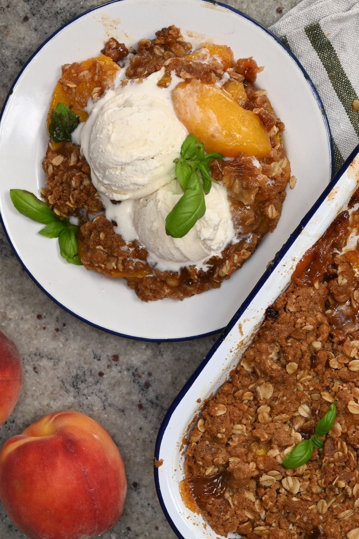 Top-down view of a serving of peach crisp topped with vanilla ice cream and basil leaves on a white enamel plate, next to two fresh whole peaches and the baking dish.